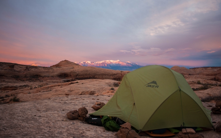 A tent rests in a desert landscape. The sky appears in shades of purple and pink, and there is a snow-capped mountain in the distance. 